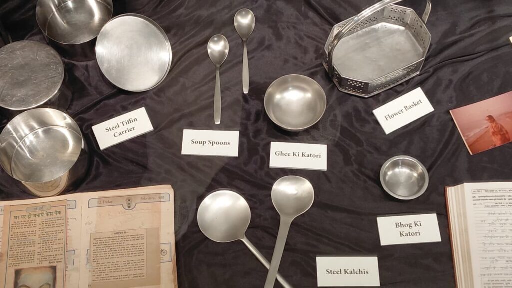 Steel tiffin carrier, soup spoons, ghee ki katori and other kitchen objects laid out with handwritten diary pages in the background